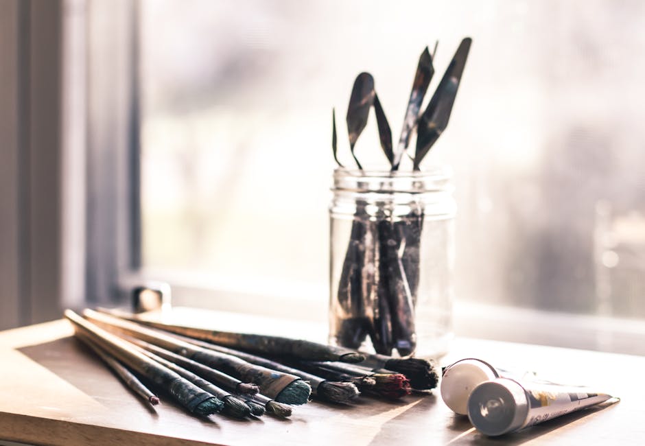 Glass jar with palette knives and paintbrushes on a sunny table, evoking creativity.
